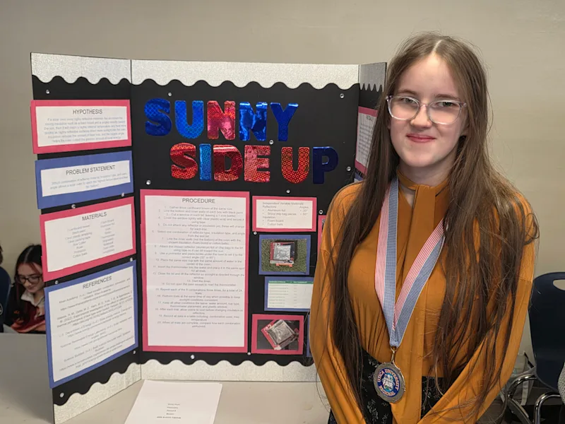 student standing with their science project, 'sunny side up', at the utica university science fair, wearing their ribbon they received