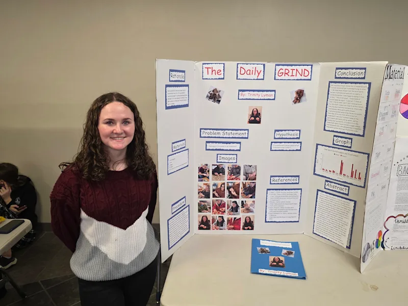 student standing next to their project, 'the daily grind', at the utica university science fair smiling