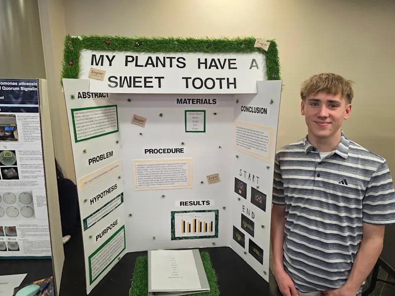 student standing next to their project, 'my plants have a sweet tooth', at the utica university science fair