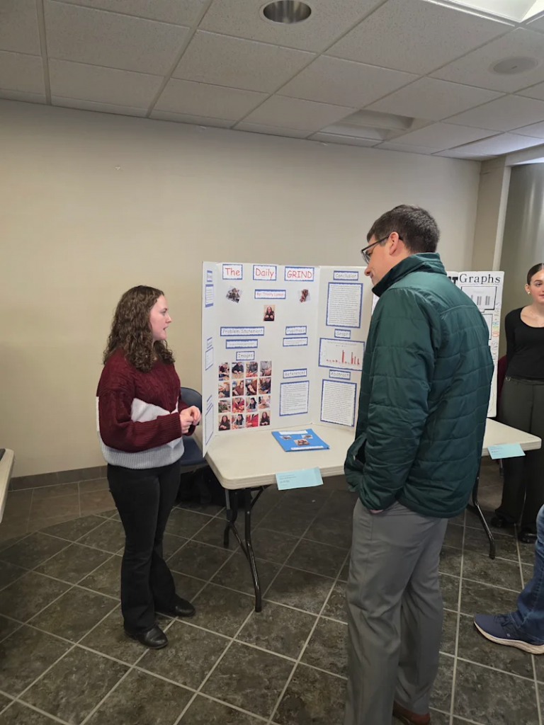 student standing next to their project, 'the daily grind', at the utica university science fair, talking to someone, explainging their idea/project
