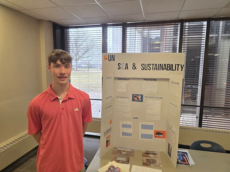 student standing next to their project, 'UN sea and sustainability', at the utica university science fair