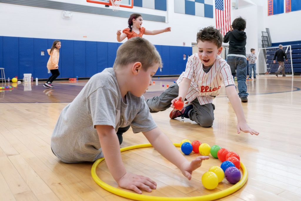 Two kids playing in gym class putting plastic balls in hula hoop